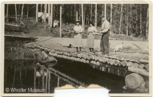 Margaret Tapley, Edna, & her husband Don McRae with dog Ki, fishing from the log bridge to Tracks, Myrtle's tent house. 1915. Inscription reads: Rainbow 1915. Philip Collection.